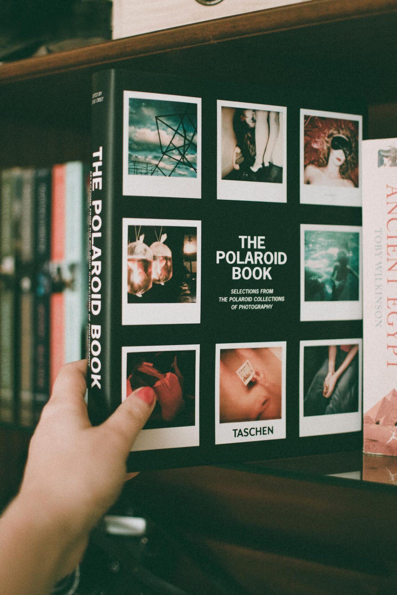 Close-up of a hand holding The Polaroid Book on a bookshelf, showcasing film photography.